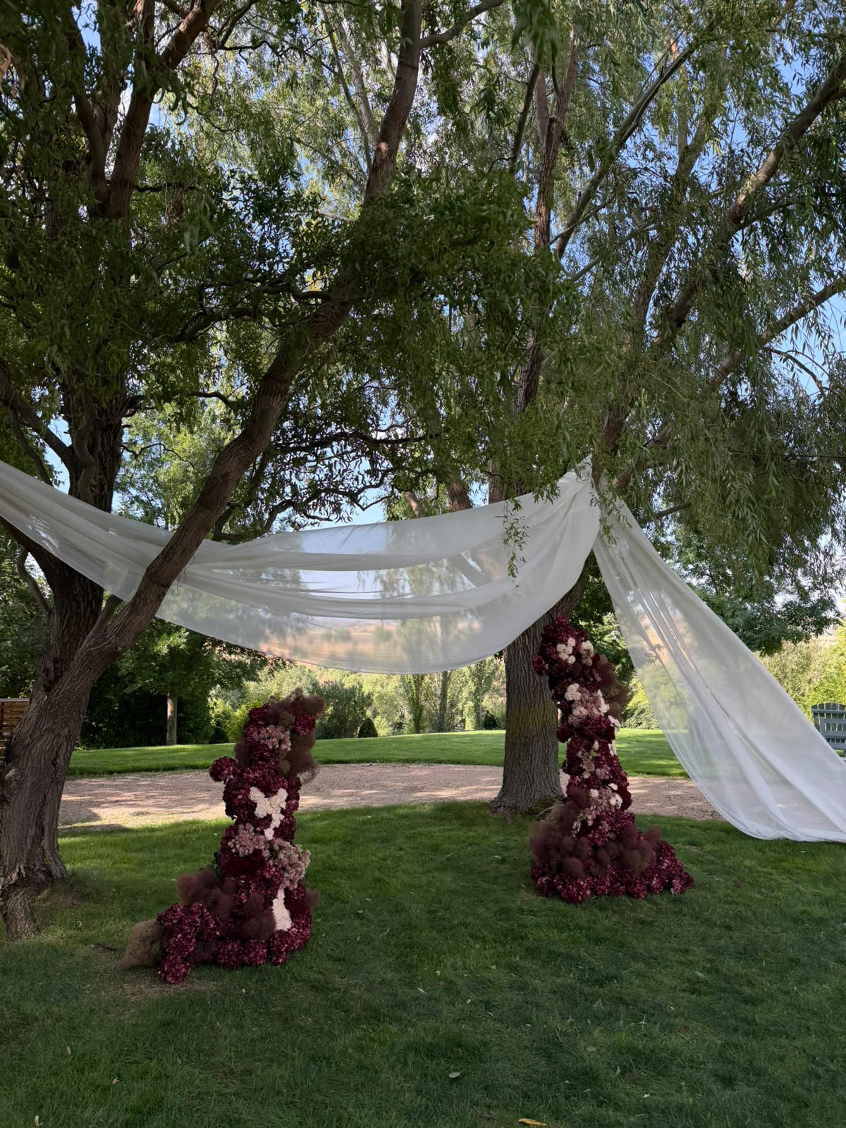 Ceremonia de boda al aire libre con columnas florales burdeos y telas blancas entre árboles